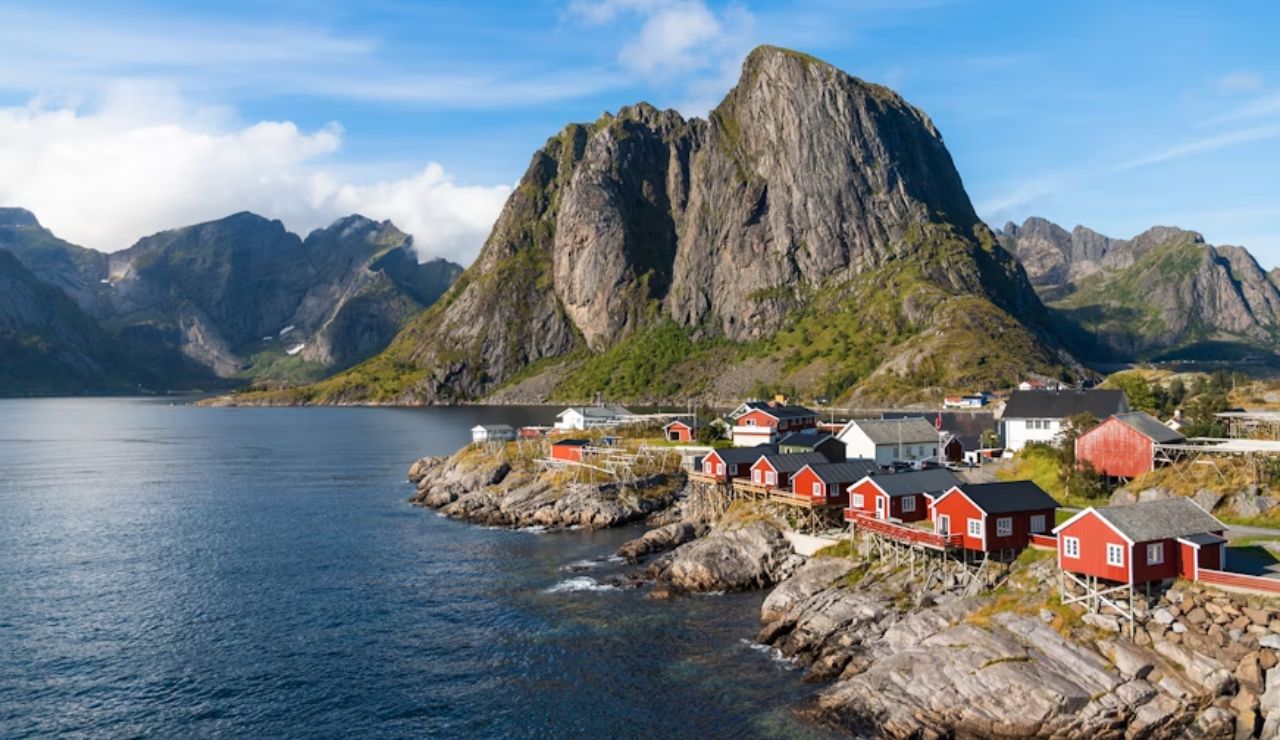 Coastal village in Lofoten, Norway, with traditional red cabins on rocky shores and dramatic mountain peaks rising in the background.