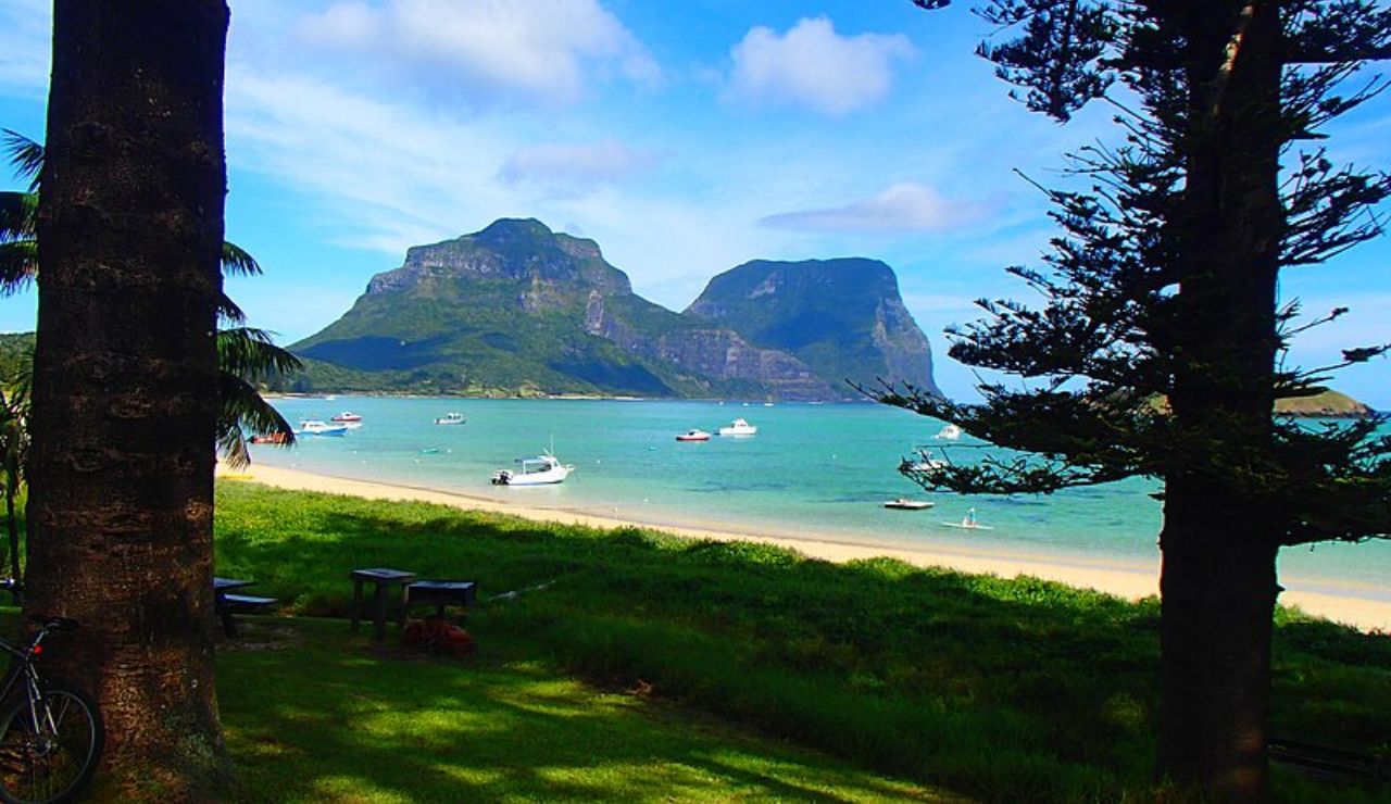 Scenic view of a beach with boats anchored in turquoise water, framed by trees and grass, with twin-peaked mountains in the background.