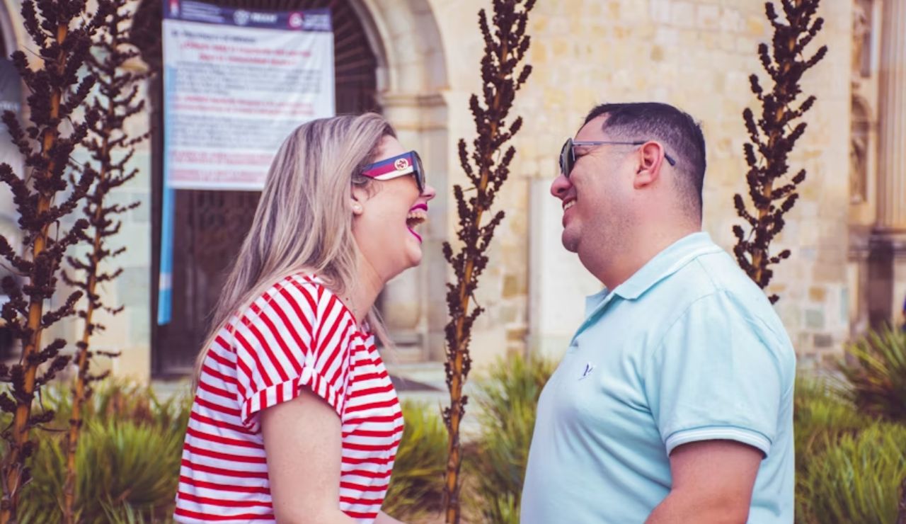 A couple wearing sunglasses, laughing and facing each other in a playful moment outdoors.