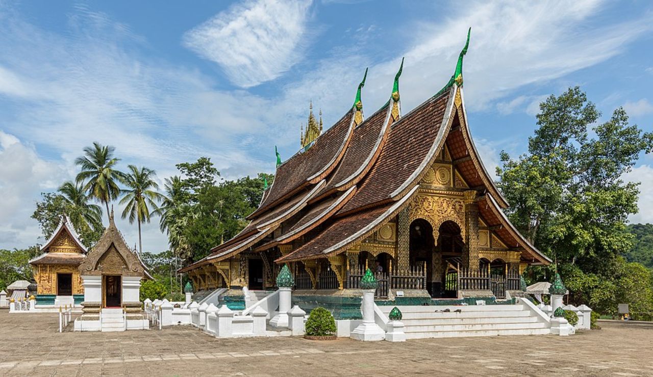 Ornate Buddhist temple with a multi-tiered roof and intricate gold detailing, surrounded by trees and palm trees in Luang Prabang, Laos.