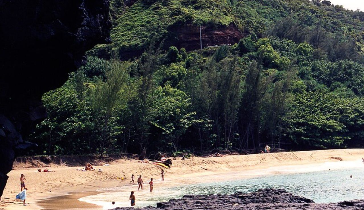 Secluded tropical beach with people enjoying the shore, surrounded by dense green forest and rocky cliffs.
