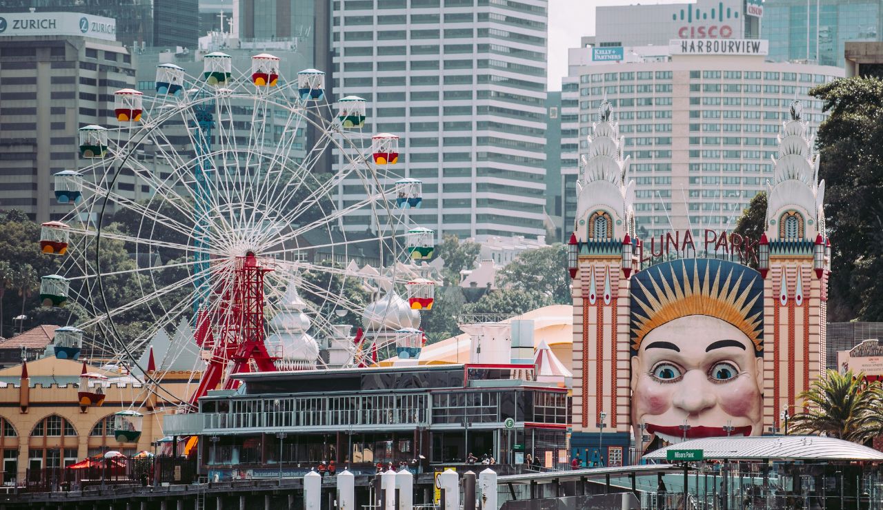 Luna Park entrance in Sydney with its iconic smiling face gate and colorful Ferris wheel, set against a backdrop of modern city buildings.
