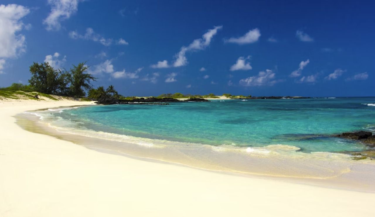 Pristine beach with soft white sand, turquoise waters, and scattered black rocks under a bright blue sky.
