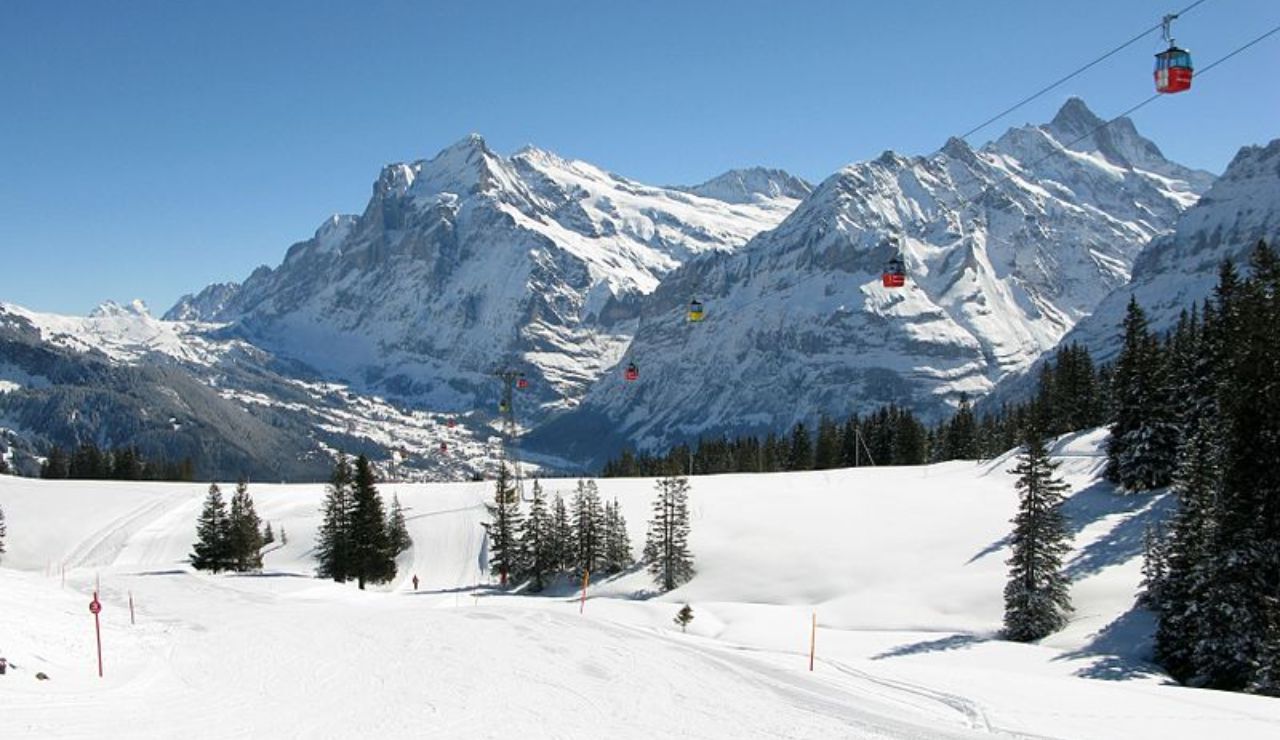 Snow-covered alpine landscape with cable cars crossing above ski slopes, surrounded by tall pine trees and majestic mountain peaks.