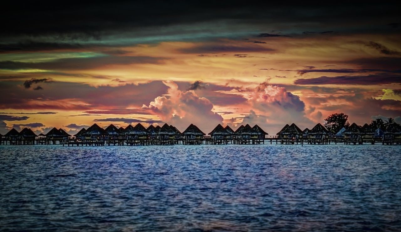 Overwater bungalows line the shore of Matira Beach in Bora Bora, set against a dramatic sunset sky and gently rippling ocean.