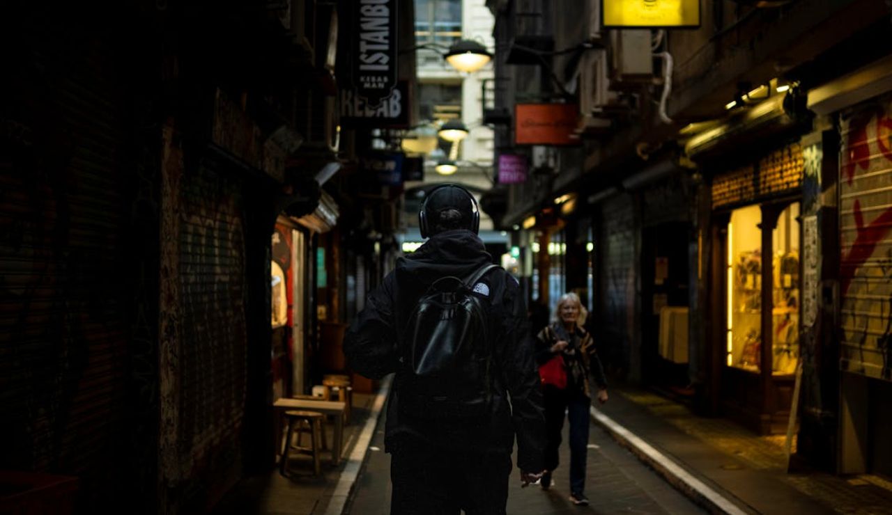 Person wearing headphones and a backpack walking through a dimly lit, narrow urban alley lined with small shops and glowing signs.