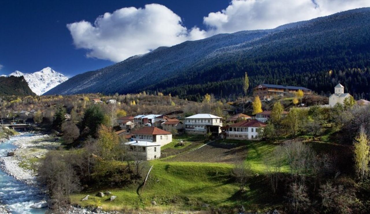 Mountain village with traditional houses, a river flowing nearby, and snow-capped peaks in the background under a partly cloudy sky.