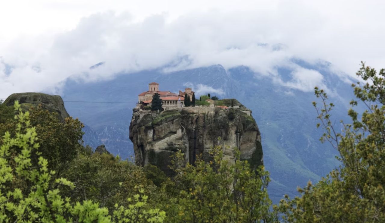 Monastery perched on top of a tall rock formation surrounded by mountains and clouds in the background.