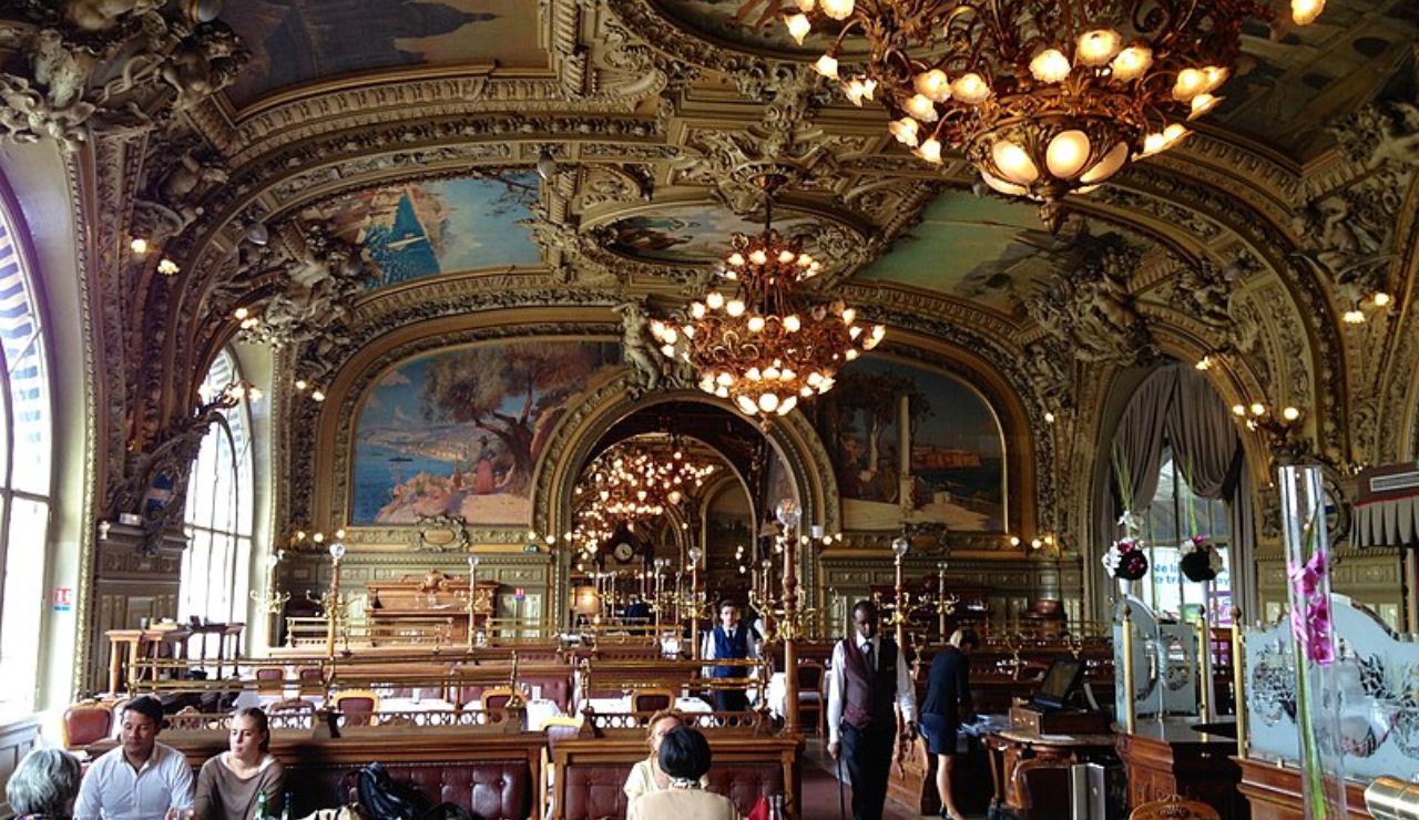 A grand, opulent restaurant interior with ornate ceilings, large chandeliers, arched windows, and classical mural paintings, filled with diners.