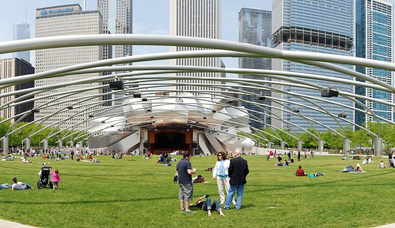 People relaxing on a large lawn at an outdoor amphitheater, surrounded by tall city buildings and overhead steel arches.
