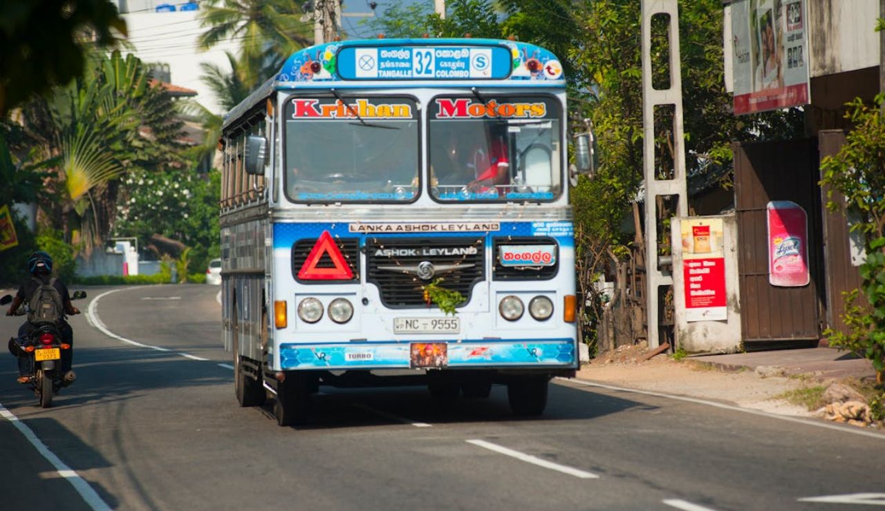 Colorful public bus labeled “Krishna Motors” driving on a sunny road with palm trees and buildings in the background.