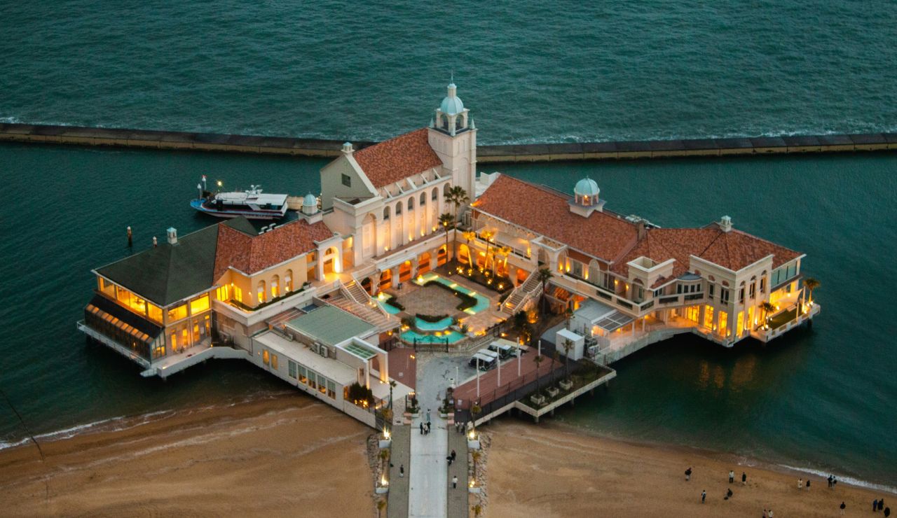 An elegant waterfront building with red-tiled roofs extends over the ocean at dusk, illuminated as people walk along a pier to its entrance.