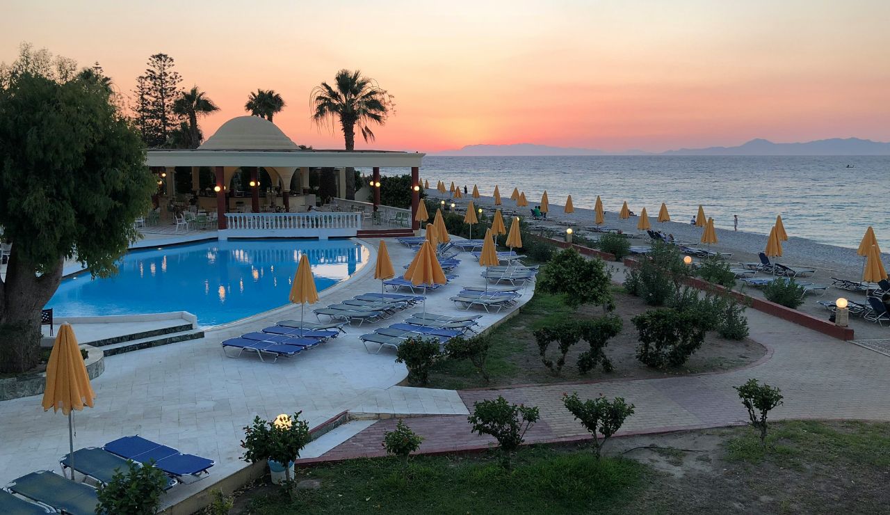 At sunset, a seaside resort features a pool with sun loungers and yellow umbrellas, overlooking the beach, calm sea, palm trees, and a pavilion.