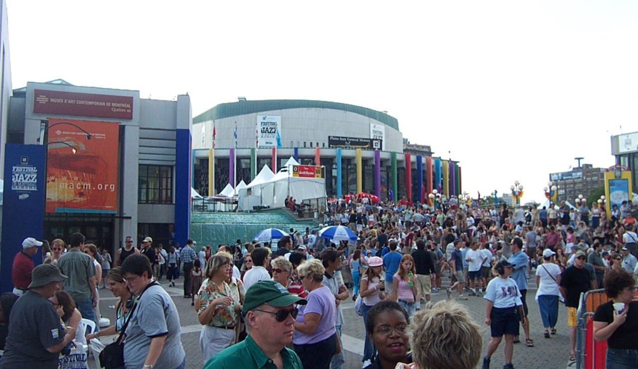 A large crowd gathers outside a colorful performing arts venue during a jazz festival, with people walking, talking, and enjoying the lively atmosphere.