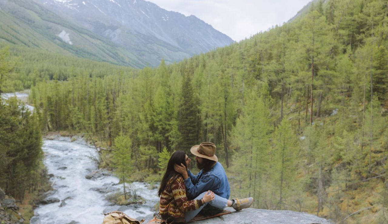 A couple sits lovingly on a rock ledge overlooking a forested valley with a rushing river, surrounded by pine trees and distant snow-capped mountains.