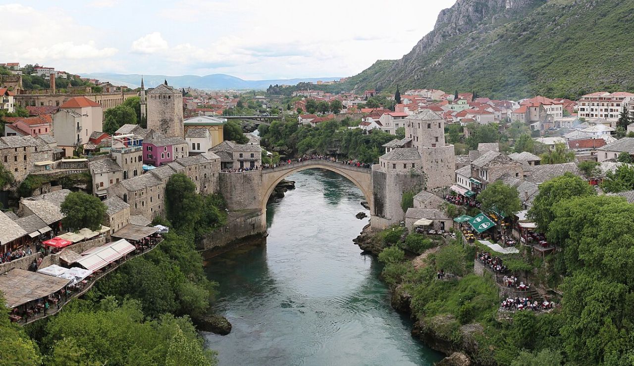 Scenic view of the historic Stari Most bridge in Mostar, Bosnia and Herzegovina, spanning a turquoise river surrounded by stone buildings and lush greenery.