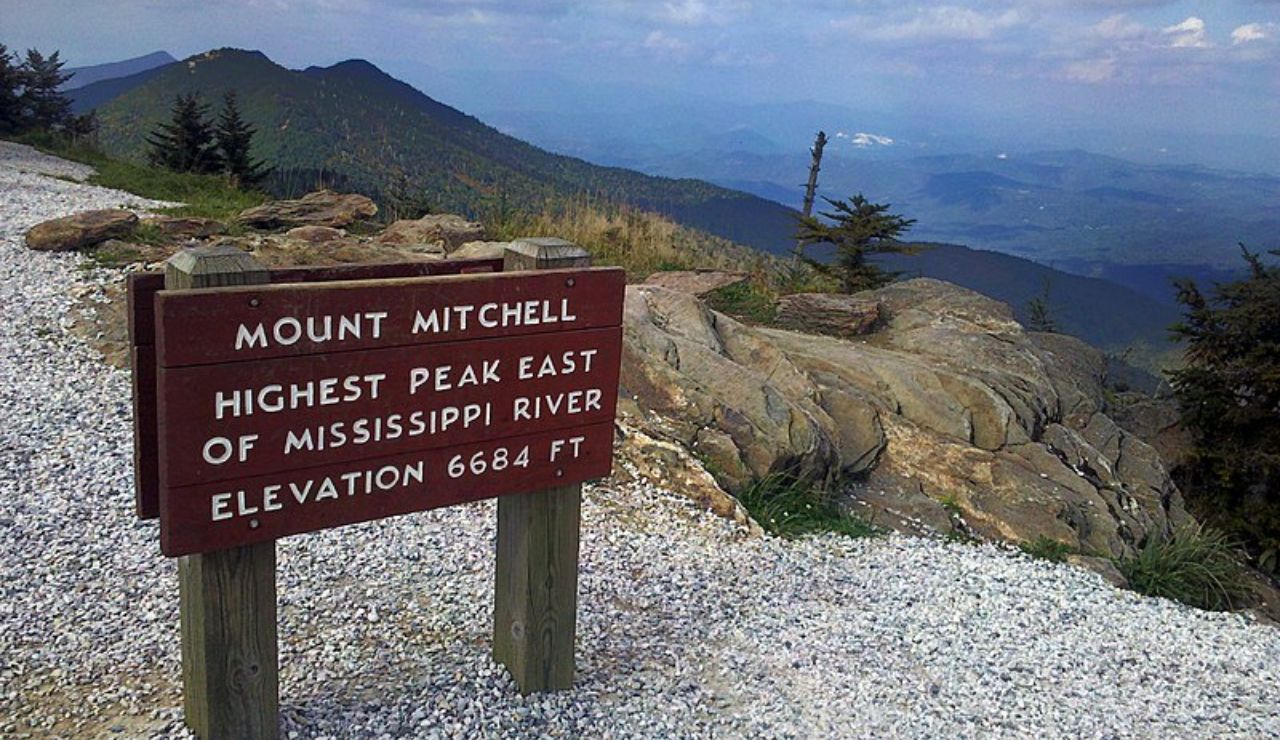 A wooden sign at Mount Mitchell marking it as the highest peak east of the Mississippi River with an elevation of 6,684 feet, overlooking a scenic mountain view.