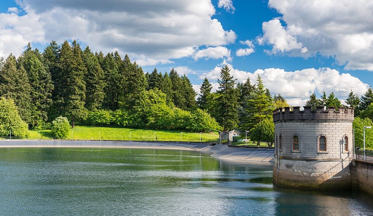 Scenic reservoir surrounded by lush green trees, with a small stone turret building along the water’s edge under a partly cloudy sky.