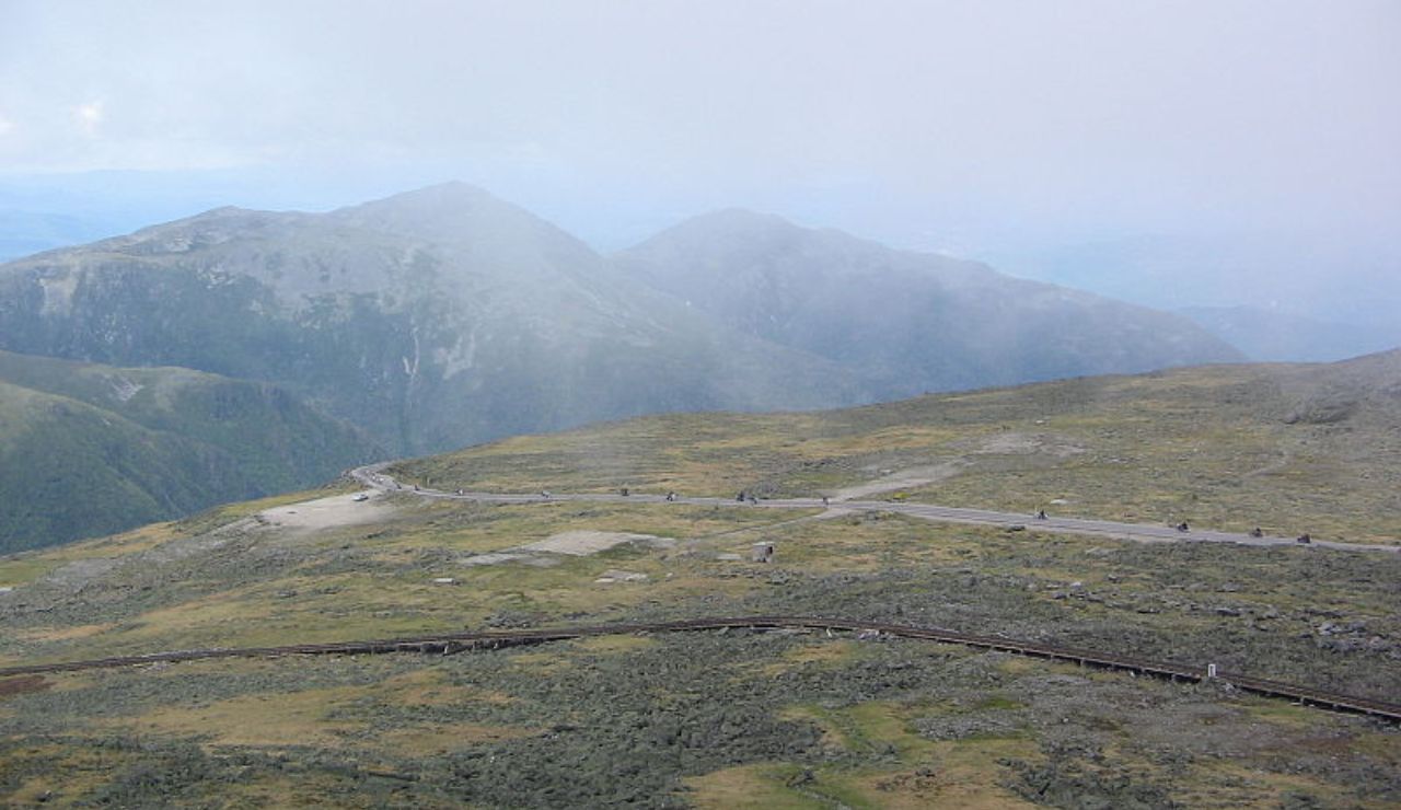 A misty mountain landscape with a winding road across a grassy plateau, leading toward hazy peaks in the distance.