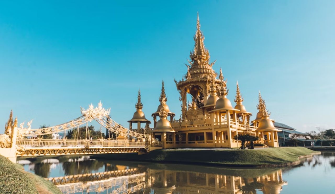 Ornate golden temple with spires and a bridge reflected in water under a clear blue sky in Thailand.