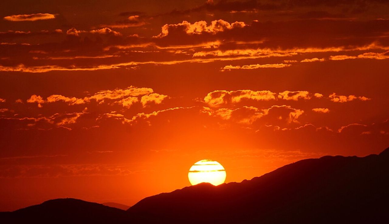 Bright orange sun setting behind a silhouette of mountain ridges with dramatic clouds in the sky.