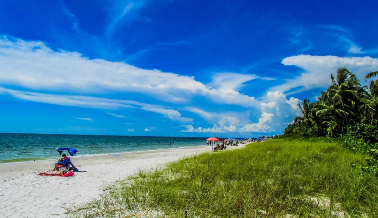 A vibrant beach scene with white sand, scattered umbrellas, lush greenery, and dramatic blue skies with swirling clouds.