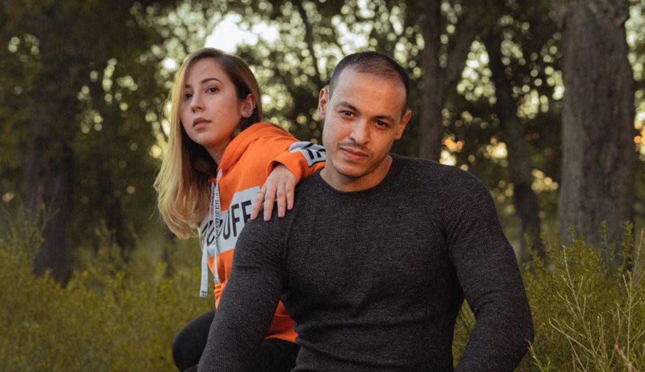 A man and a woman posing together in a forest setting, with the woman standing behind the man, both looking at the camera.