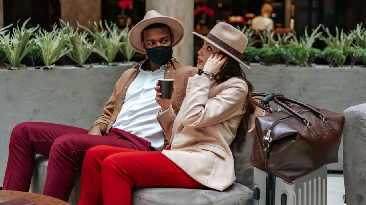 A stylish couple sits together in a lounge area with luggage, wearing coordinated outfits and hats; the man wears a face mask.