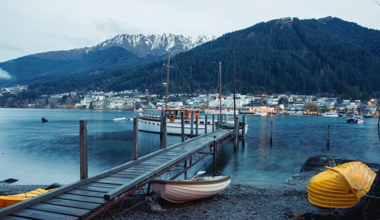Wooden dock leading to a sailboat on a calm lake, with snow-capped mountains and a lakeside town in the background.