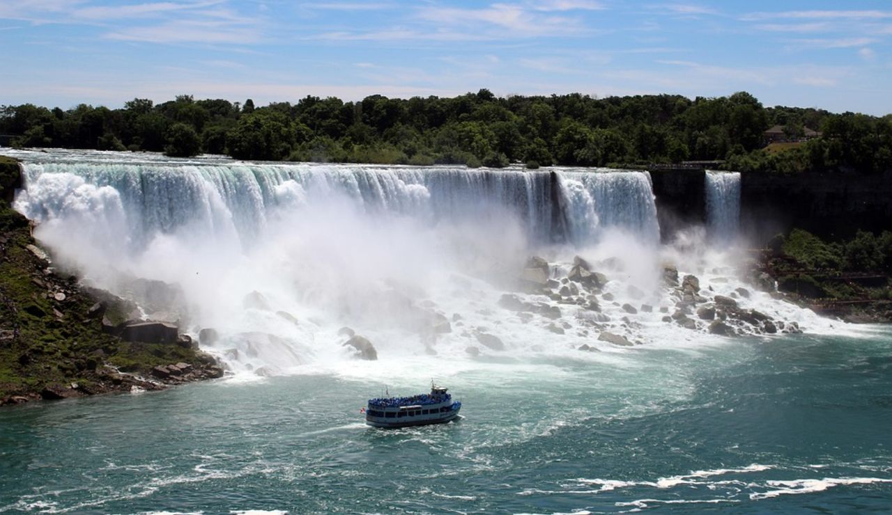 Powerful waterfall cascading into a misty river as a tour boat approaches below.