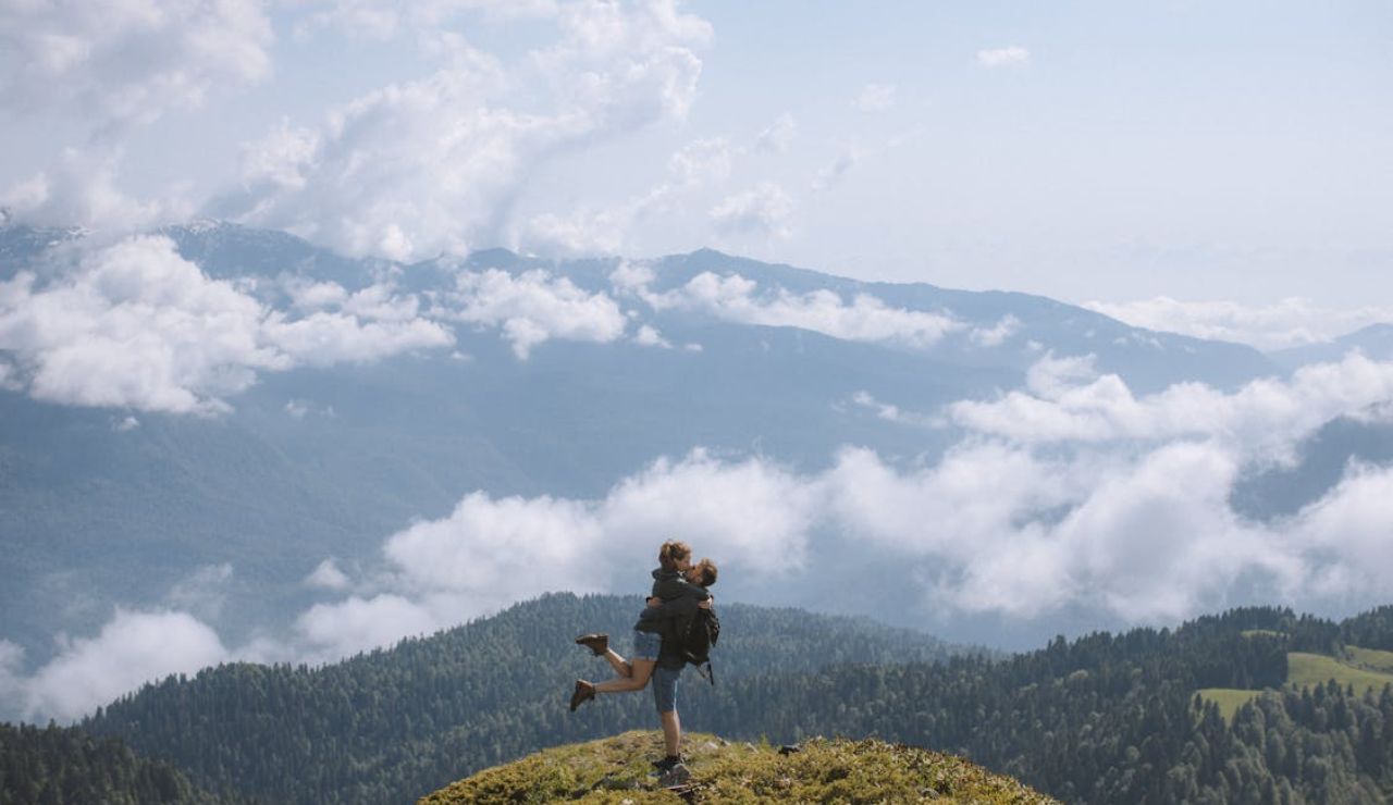 A joyful couple embraces on a mountain peak surrounded by clouds and vast forested hills under a bright, partly cloudy sky.