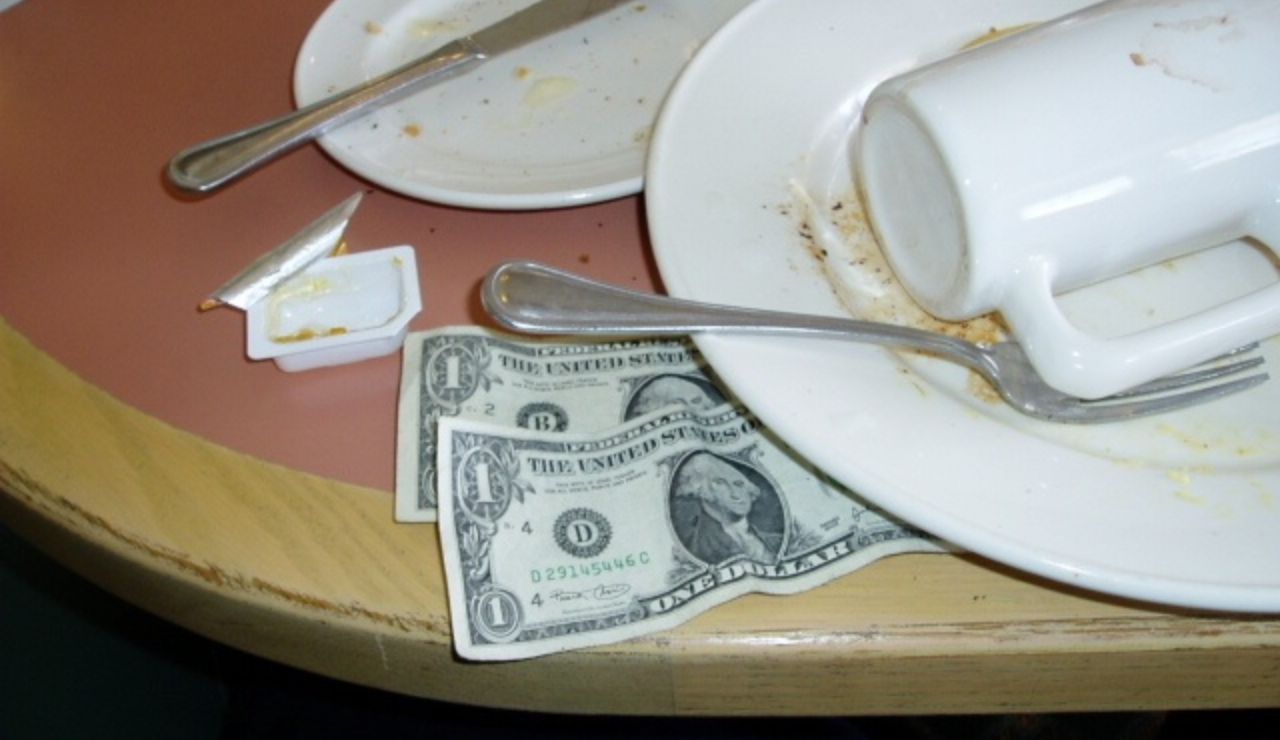 Two one-dollar bills left as a tip on a diner table with dirty dishes, utensils, and an overturned coffee mug.