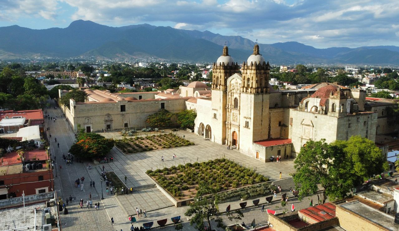 Aerial view of a historic cathedral with twin bell towers and a central courtyard garden, set against a backdrop of a city and distant mountains.