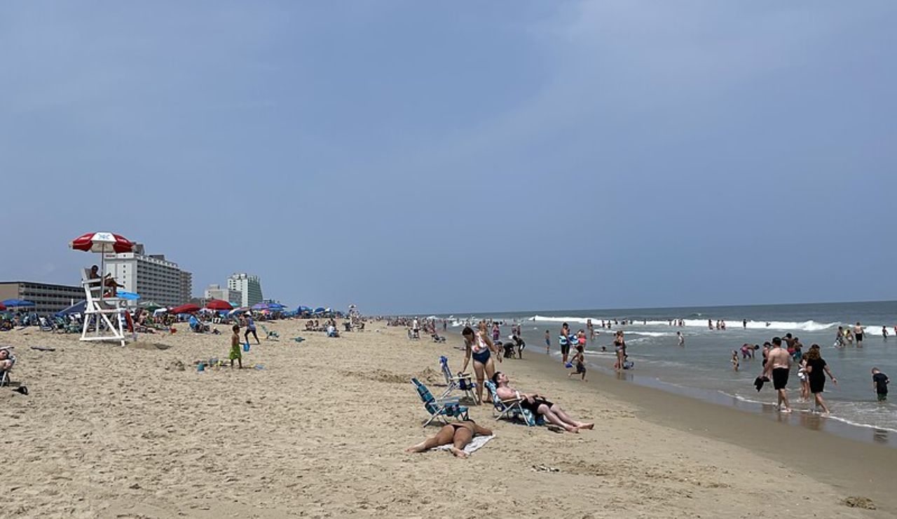 Crowded beach with people sunbathing, swimming, and walking along the shore near tall beachfront buildings and a lifeguard stand.