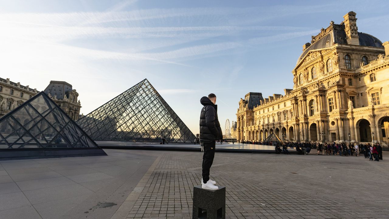 A person in a black jacket stands on a pedestal near the Louvre pyramids, with museum buildings and a crowd in the background.