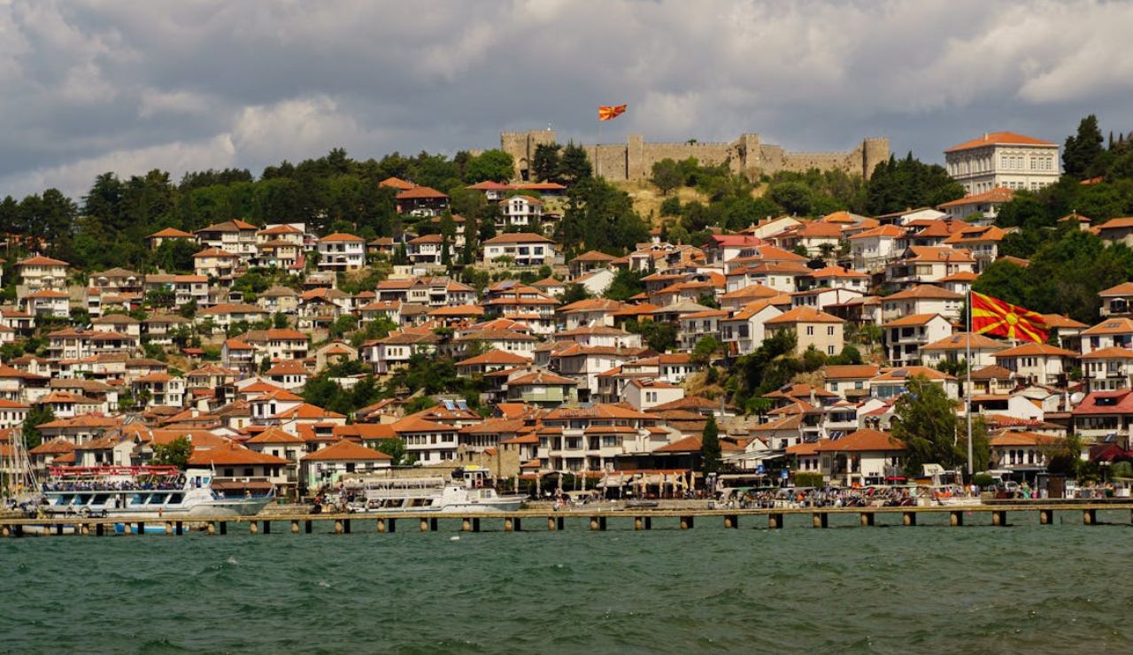 Lakeside view of Ohrid, North Macedonia, with red-roofed houses, a historic fortress on the hill, and Macedonian flags flying above the town.