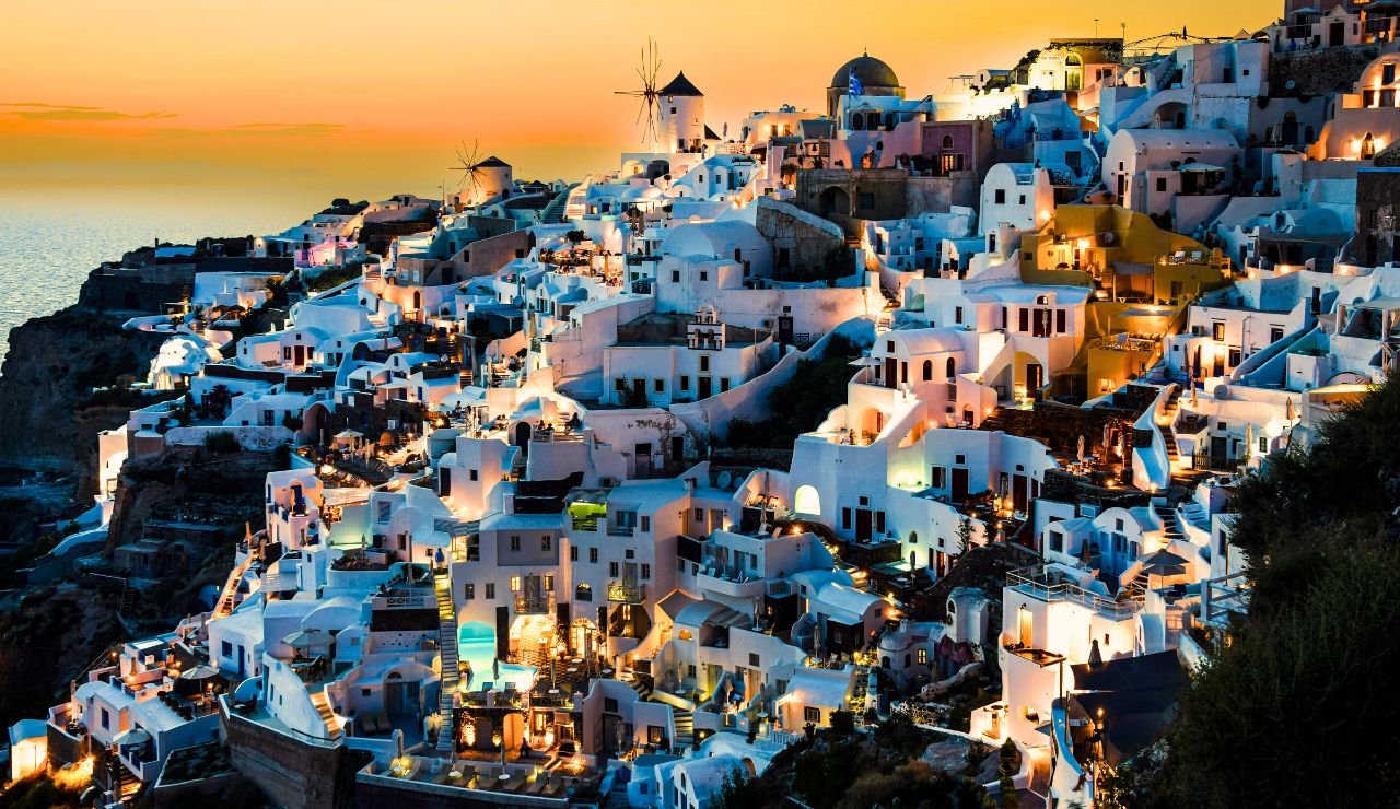 At sunset, Santorini's white-washed buildings and windmills glow above the sea against a vibrant orange sky.