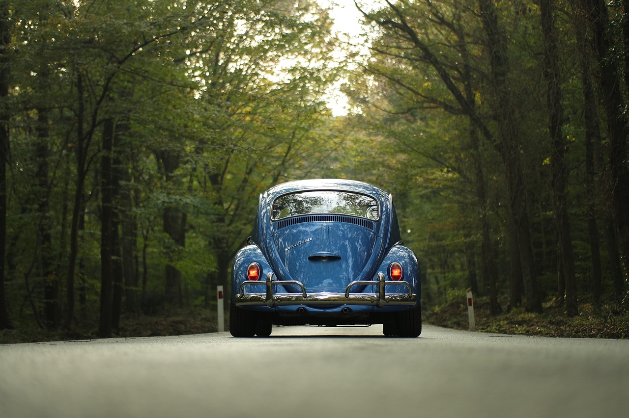 A blue vintage beetle car drives down a paved road through a forest.