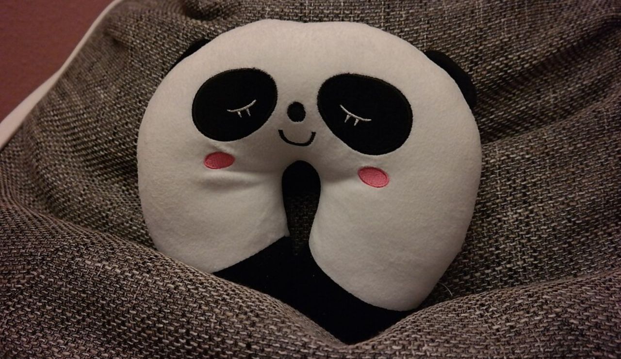 A cute panda-themed U-shaped neck pillow with closed eyes and pink cheeks, resting on a textured gray blanket.