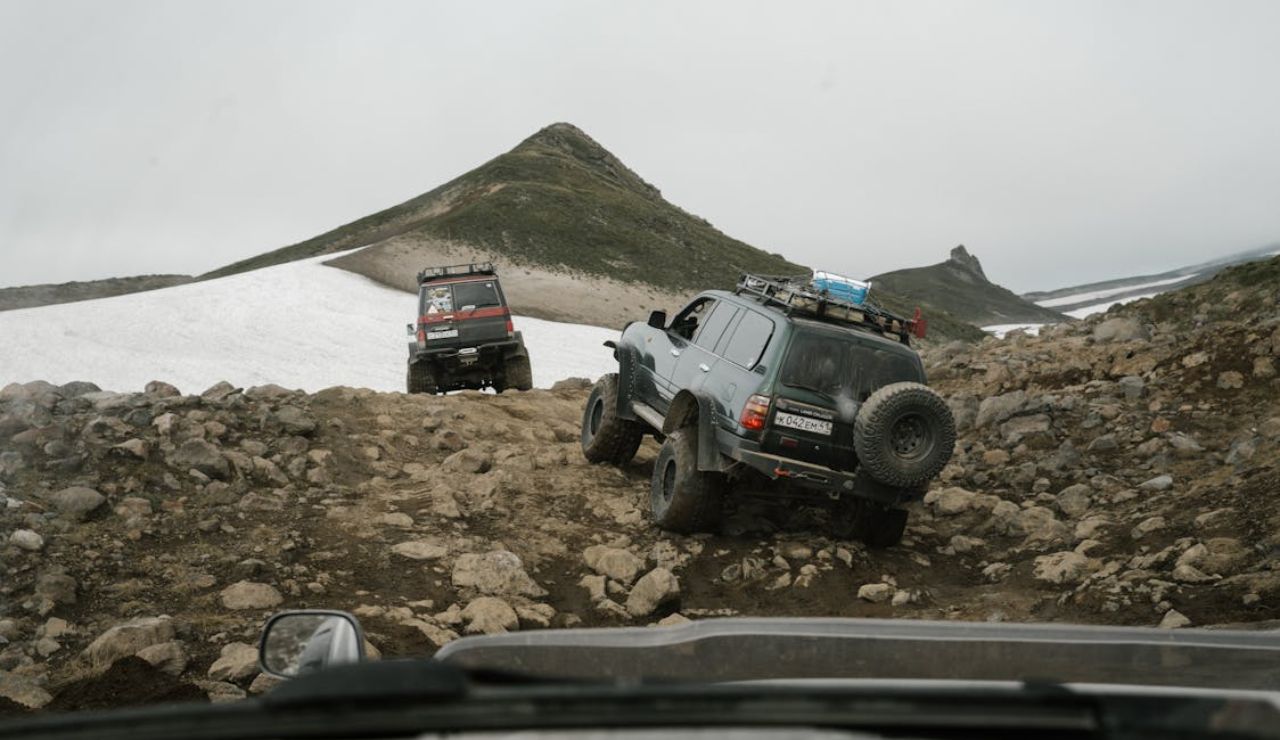 Off-road vehicles driving on a rocky mountain trail with patches of snow and rugged peaks in the background.