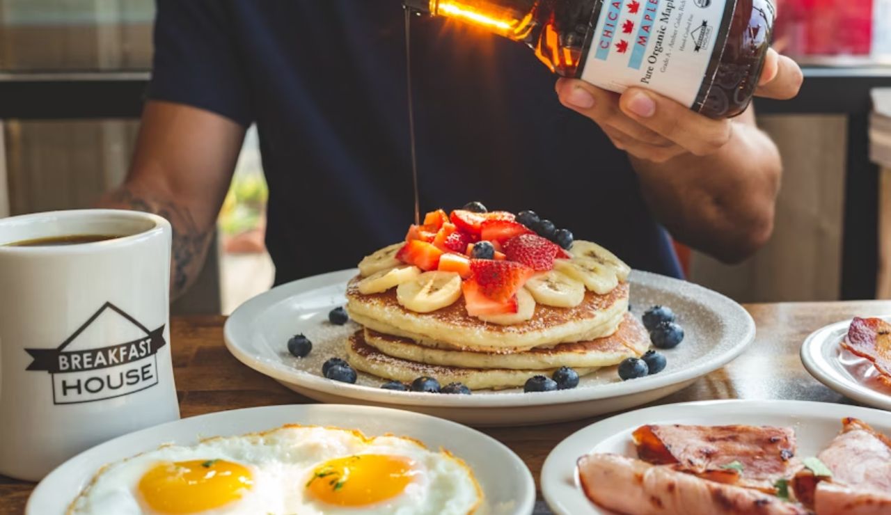 A person pouring maple syrup over a stack of pancakes topped with fresh fruit, accompanied by eggs, bacon, and a cup of coffee labeled "Breakfast House."