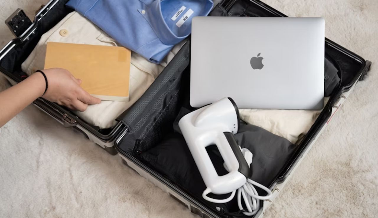 Open suitcase on a carpeted floor with folded clothes, a laptop, a handheld garment steamer, and a person placing a book inside.