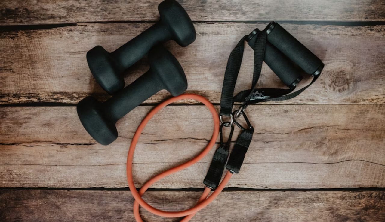 Two black dumbbells and an orange resistance band with handles placed on a wooden floor.