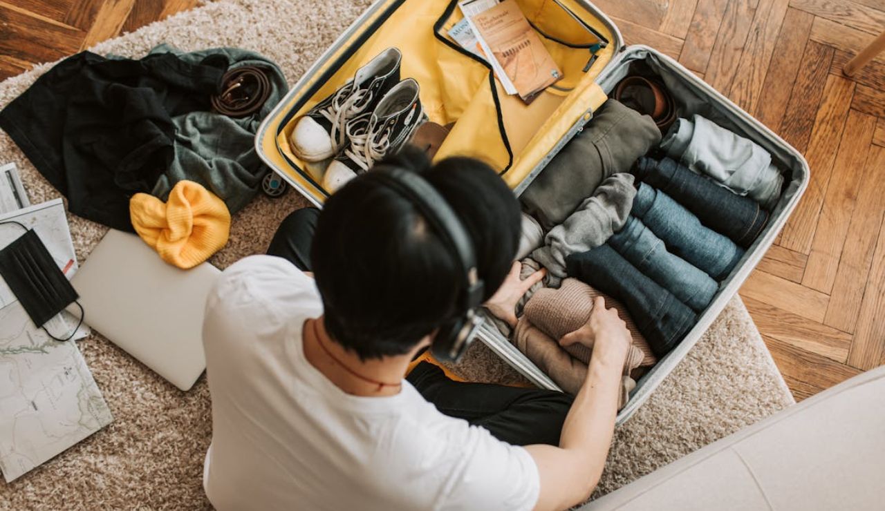 Overhead view of a person packing neatly folded clothes and shoes into a suitcase, with travel essentials scattered nearby.