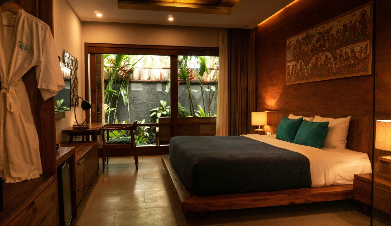 A modern hotel room features a large bed, wooden furniture, wall art, a window desk, lamps, and garden views through sliding glass doors.