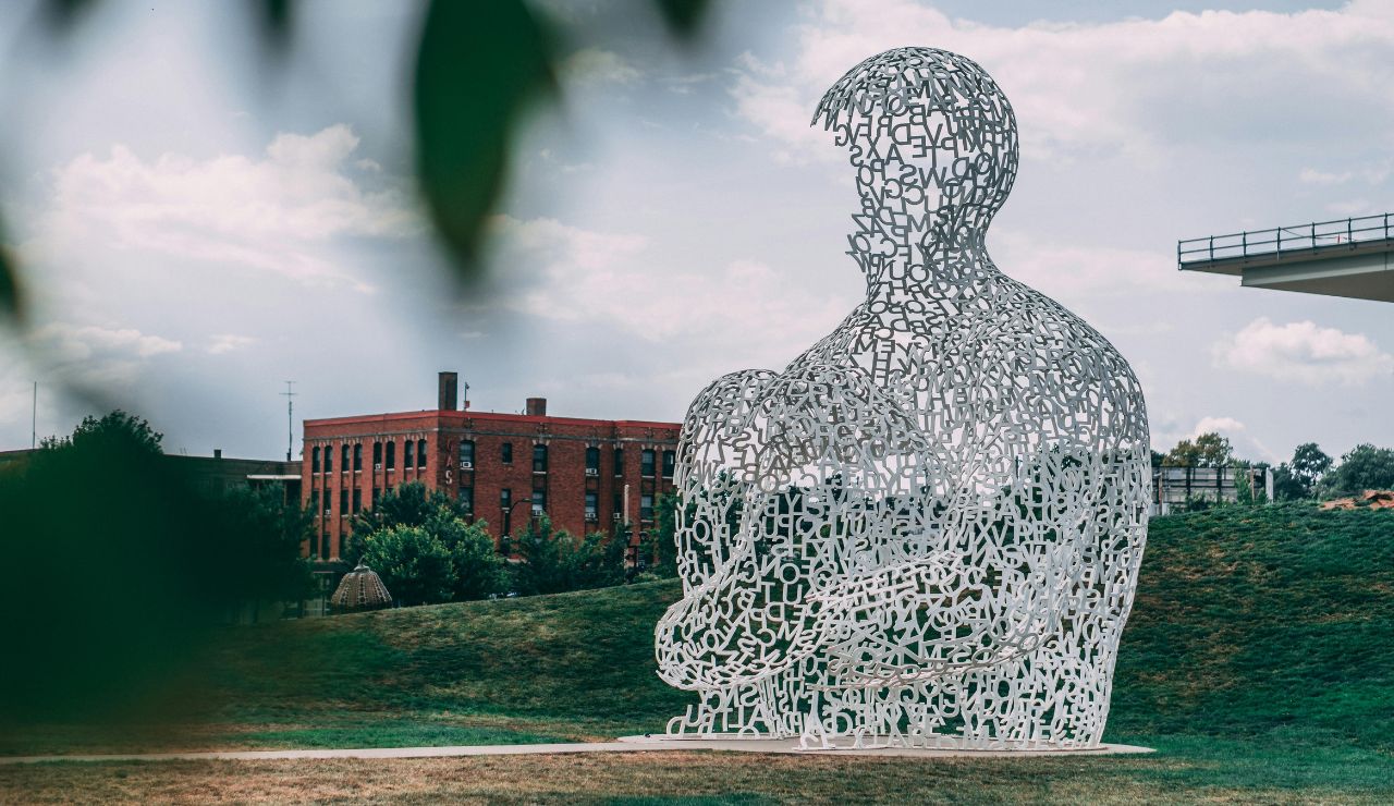 Large outdoor sculpture of a seated human figure made from interlocking letters sits on a grassy hill near a brick building, with trees and a cloudy sky behind.