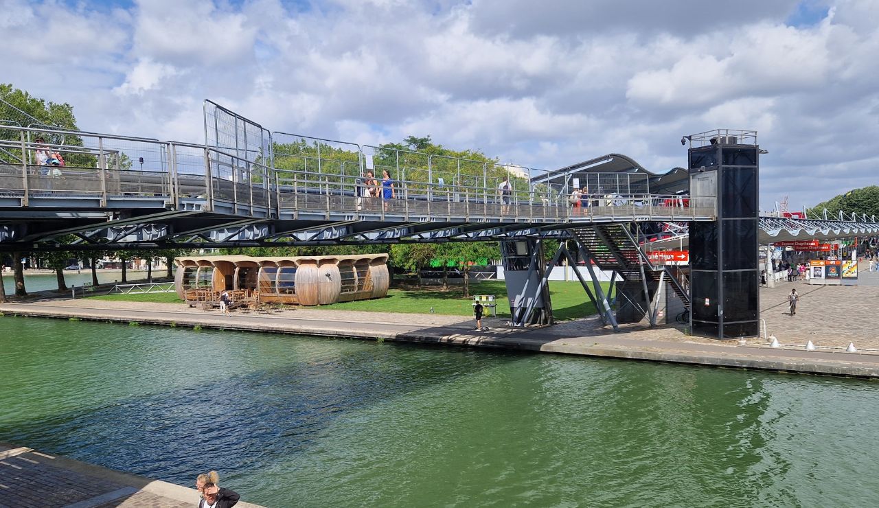 People walk on a metal pedestrian bridge over a green canal and along its edge beneath a partly cloudy sky.