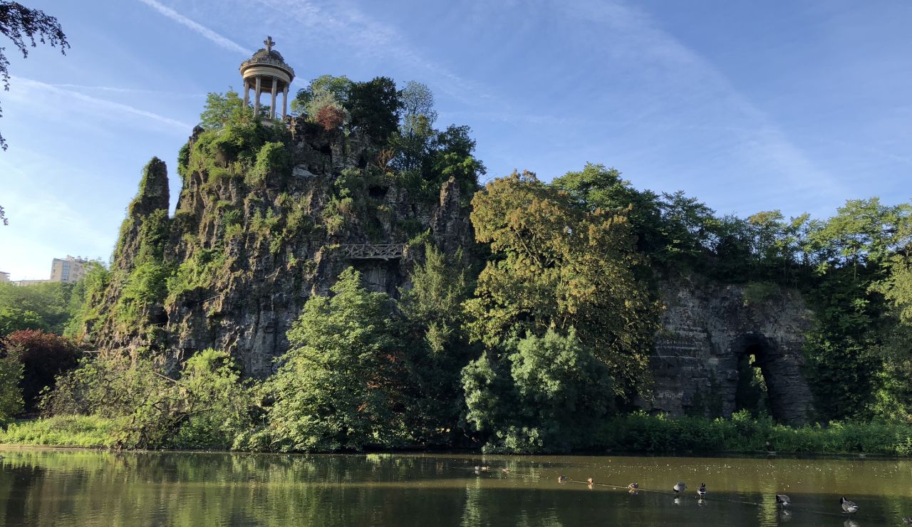 A rocky hill topped with a classical pavilion overlooks a tree-lined pond where ducks swim beneath a clear blue sky.