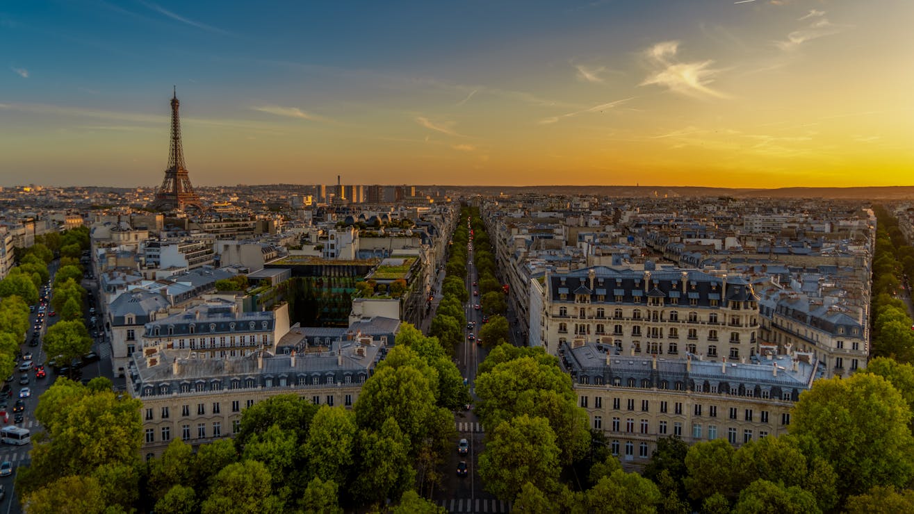 Aerial view of Paris at sunset with the Eiffel Tower in the distance, tree-lined avenues, and classic Haussmann-style buildings.