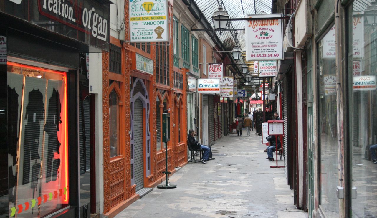 A narrow indoor market corridor features various shop and restaurant signs, people sitting on benches, and a glass roof overhead.